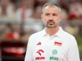 Skupiony trener siatkarzy reprezentacji Polski w białej koszulce z godłem narodowym i logotypami sponsorów.