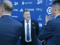 Mężczyzna w garniturze uśmiecha się, stojąc na tle niebieskiej ściany z logotypami sponsorów, być może rozmawiając o tym, ile dostają kluby ekstraklasy.