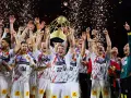 Drużyna świętuje zwycięstwo w finale piłki ręcznej, unosząc puchar. Konfetti i fajerwerki podkreślają radość.