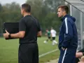 Sędzia z tablicą zmian i trener na tle meczu piłki nożnej.
