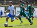 Piłkarz w białej koszulce z niebieskimi spodenkami prowadzi piłkę na boisku. W tle zawodnicy w zielonych strojach. Mecz forbet 4 liga warmińsko-mazurska.