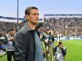 Trener Interu wpatrzony w murawę, otoczony przez fotoreporterów na tle trybun pełnych kibiców.