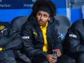 Piłkarz Adeyemi w czarnej kurtce z żółtym logo BVB i napisem REWE, siedzi na ławce rezerwowych, trzymając butelkę z napojem.