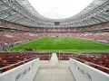 Pusta trybuna moskiewskiego stadionu z rzędem czerwonych i brązowych siedzeń. Na boisku pracują zraszacze.