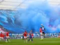 Mecz Ruch Chorzów – Wisła Kraków. Niebieskie dymy i ogromna oprawa kibiców na trybunach. Piłkarze walczą o piłkę na murawie.