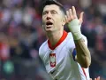 Robert Lewandowski w koszulce reprezentacji Polski, z bandażem na ręce, wznosi gest ku górze. Fani zastanawiają się, o której mecz piłki nożnej.