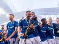 Piłkarze w niebieskich strojach świętują z pucharem i piłką na stadionie.
