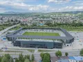 Nowoczesny stadion Sandecji z zieloną murawą i trybunami, otoczony miastem i wzgórzami.