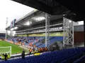 Puste trybuny stadionu Selhurst Park, z niebieskimi i czerwonymi siedzeniami, zieloną murawą i wielkim telebimem.