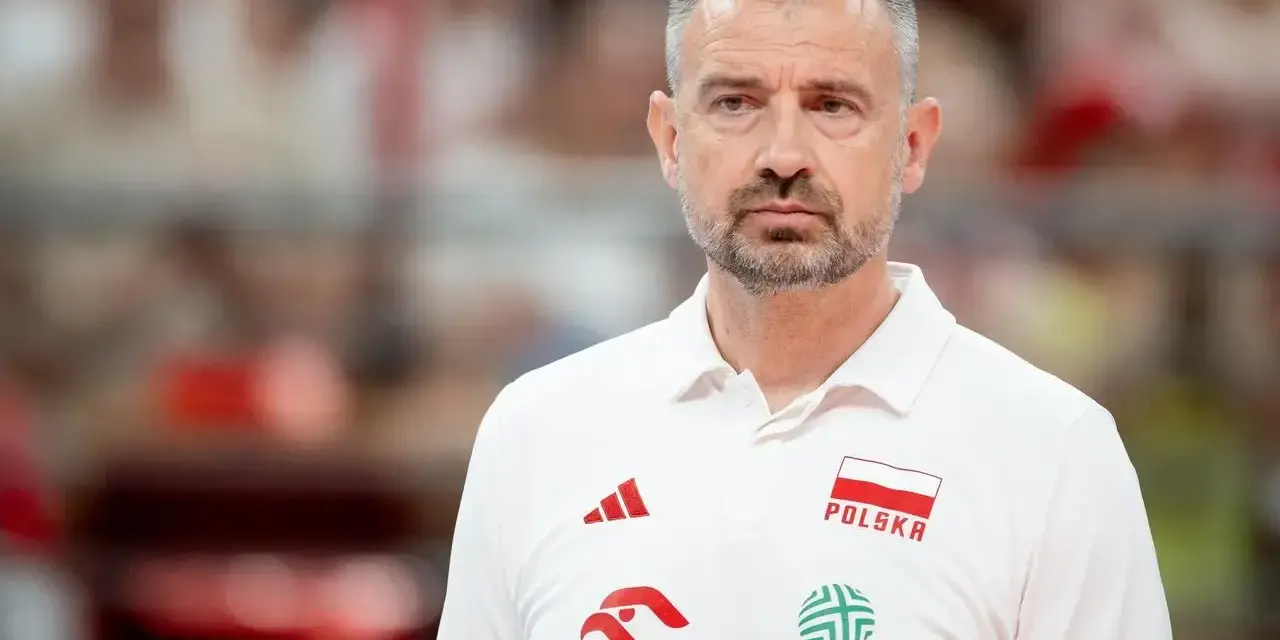 Skupiony trener siatkarzy reprezentacji Polski w białej koszulce z godłem narodowym i logotypami sponsorów.