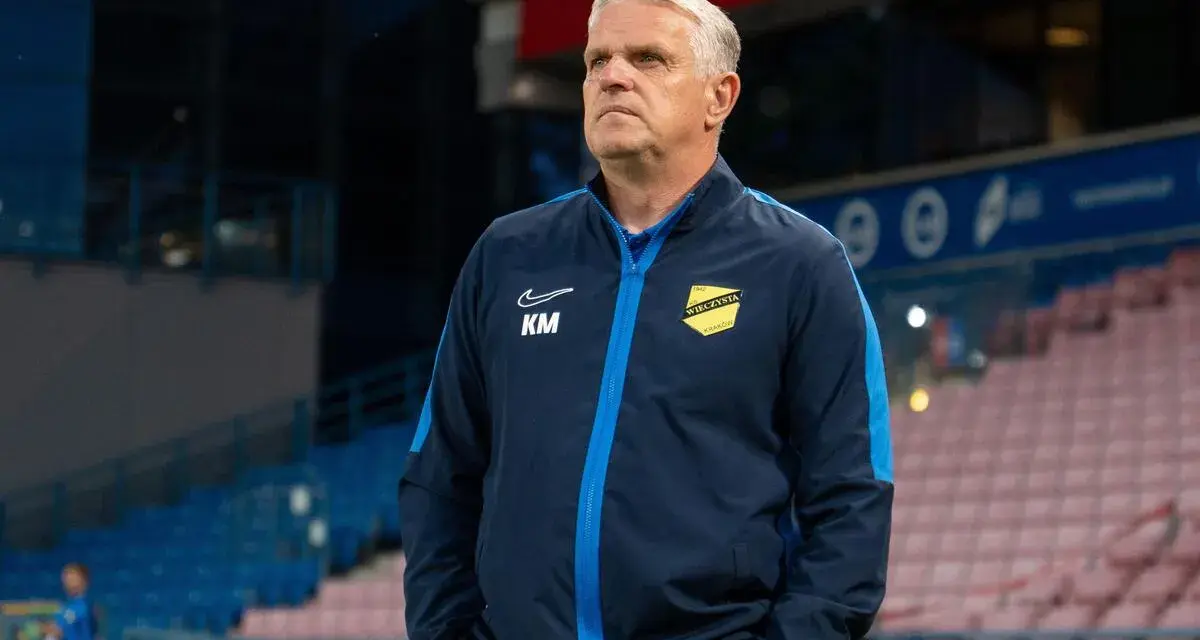 Trener z Krakowa, w granatowej kurtce z żółtym herbem, patrzy w dal na stadionie.