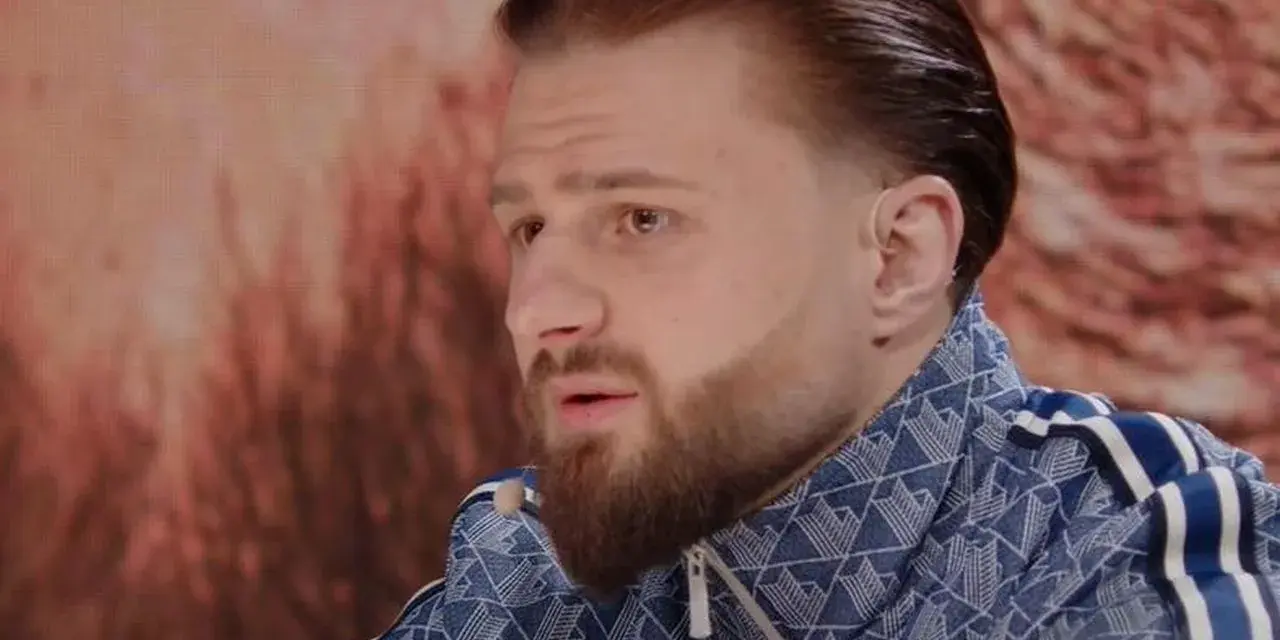 Amadeusz Ferrari w niebieskiej bluzie z białymi paskami, z charakterystyczną fryzurą i brodą, siedzi przed ekranem.