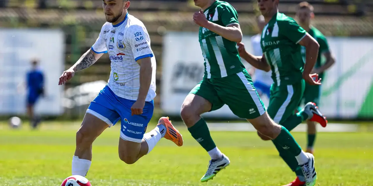 Piłkarz w białej koszulce z niebieskimi spodenkami prowadzi piłkę na boisku. W tle zawodnicy w zielonych strojach. Mecz forbet 4 liga warmińsko-mazurska.