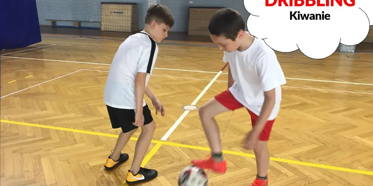 Dwóch chłopców ćwiczy drybling piłką nożną na sali gimnastycznej.