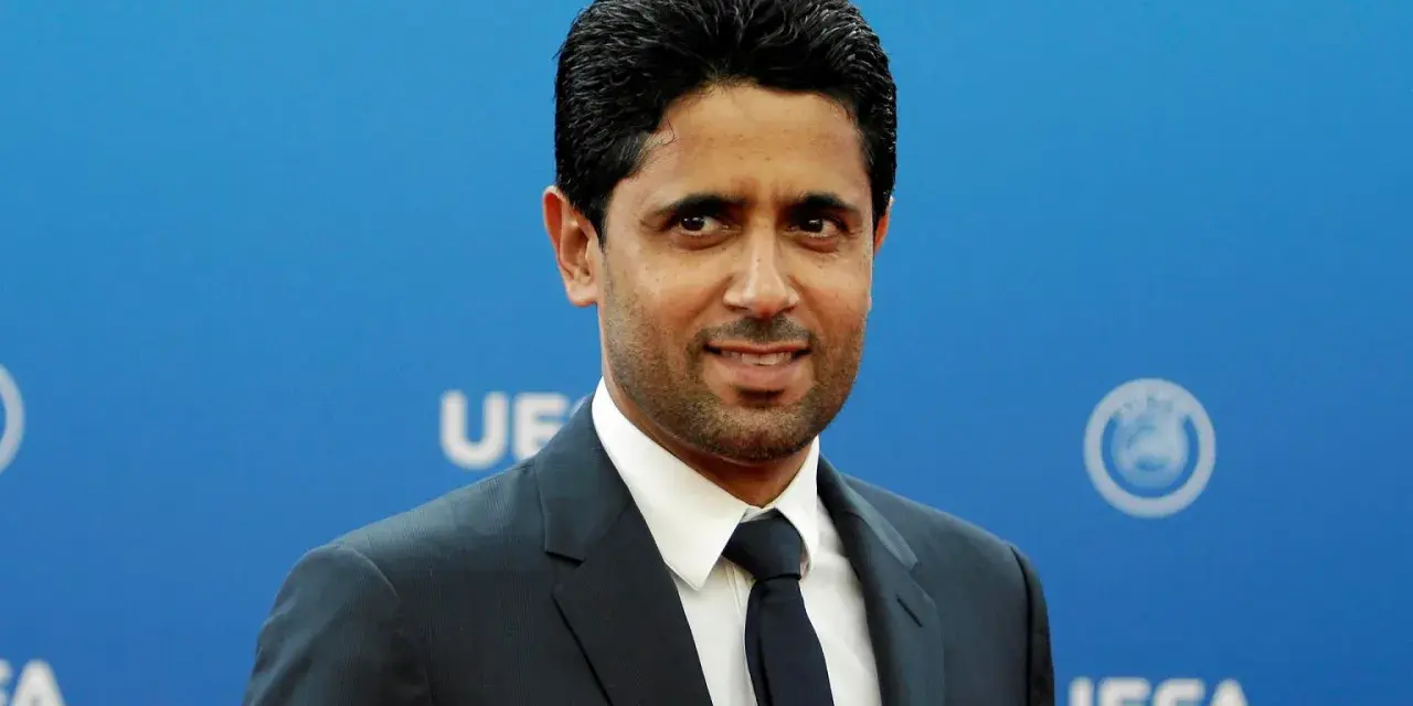 Nasser Al-Khelaifi w garniturze na tle niebieskiej ściany z logo UEFA.