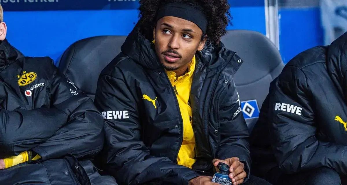 Piłkarz Adeyemi w czarnej kurtce z żółtym logo BVB i napisem REWE, siedzi na ławce rezerwowych, trzymając butelkę z napojem.