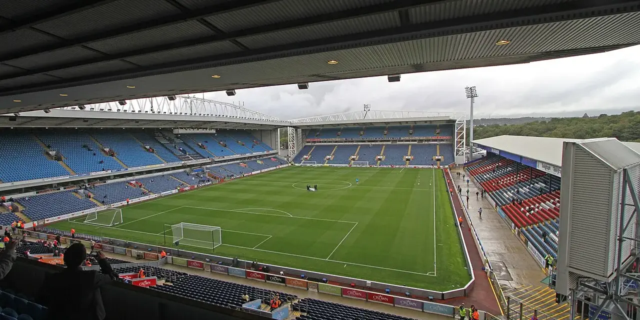Widok na murawę i trybuny stadionu Ewood Park. Puste miejsca na niebieskich i czerwonych siedzeniach.