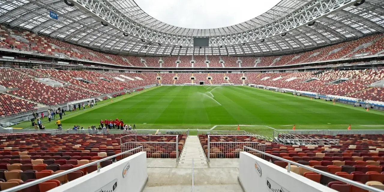 Pusta trybuna moskiewskiego stadionu z rzędem czerwonych i brązowych siedzeń. Na boisku pracują zraszacze.