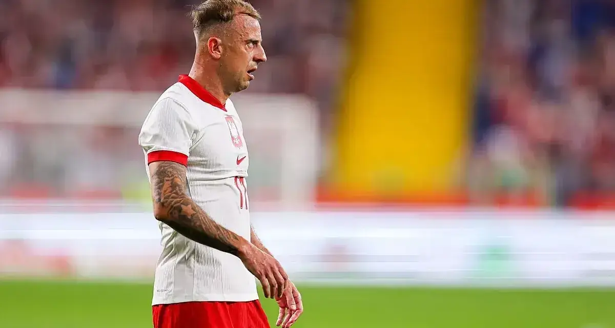 Kamil Grosicki w białej koszulce z orzełkiem i czerwonych spodenkach, gotowy do gry. Jego obecne drużyny to Polska i Pogoń Szczecin.