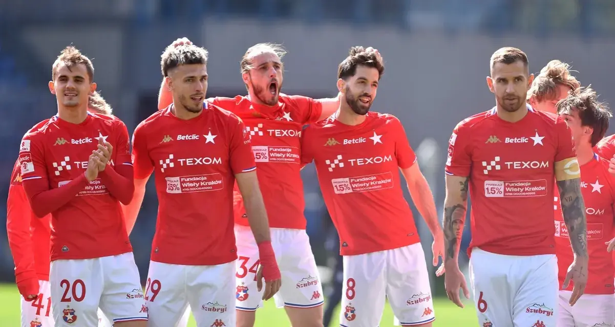 Piłkarze Wisły Kraków w czerwonych strojach, z numerami na koszulkach i spodenkach, świętują. W tle trybuny. #wisla #krakow #transfery