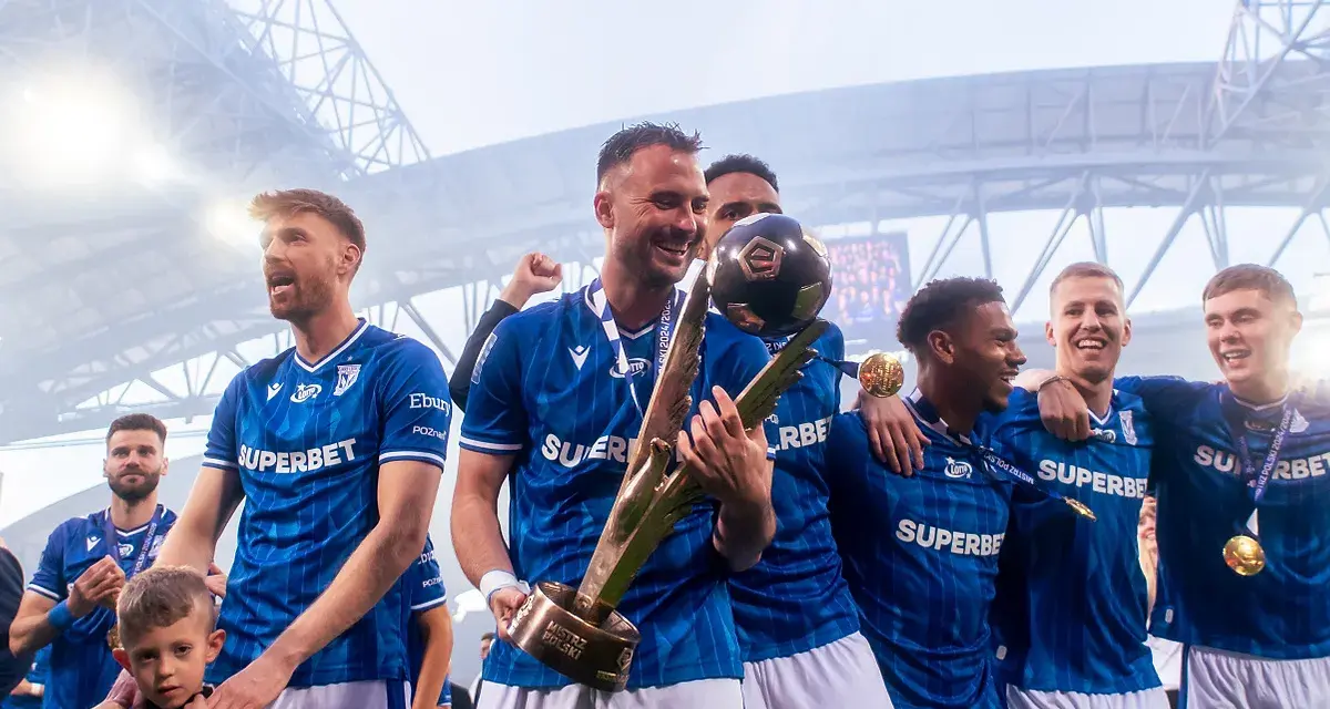 Piłkarze w niebieskich strojach świętują z pucharem i piłką na stadionie.