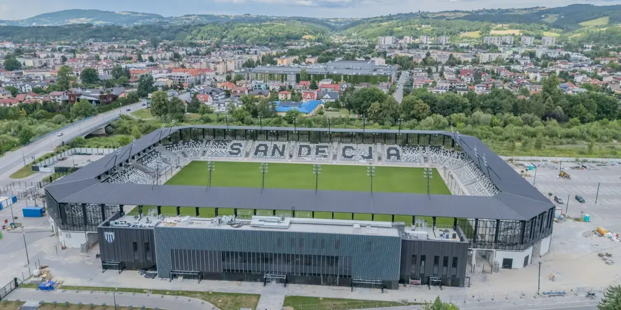 Nowoczesny stadion Sandecji z zieloną murawą i trybunami, otoczony miastem i wzgórzami.