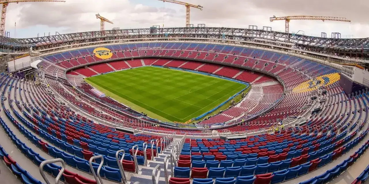 Widok na budowę stadionu, gdzie powstaje nowe Camp Nou. Żurawie górują nad trybunami w kolorach czerwonym i niebieskim, a murawa lśni zielenią.