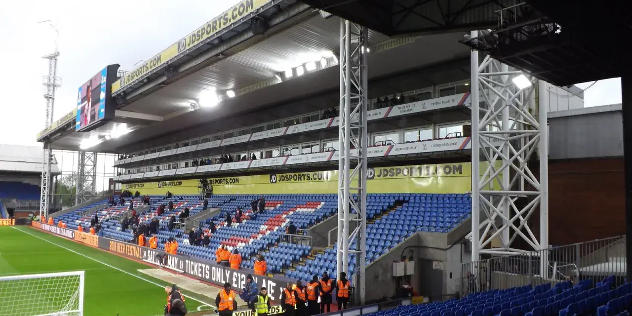 Puste trybuny stadionu Selhurst Park, z niebieskimi i czerwonymi siedzeniami, zieloną murawą i wielkim telebimem.