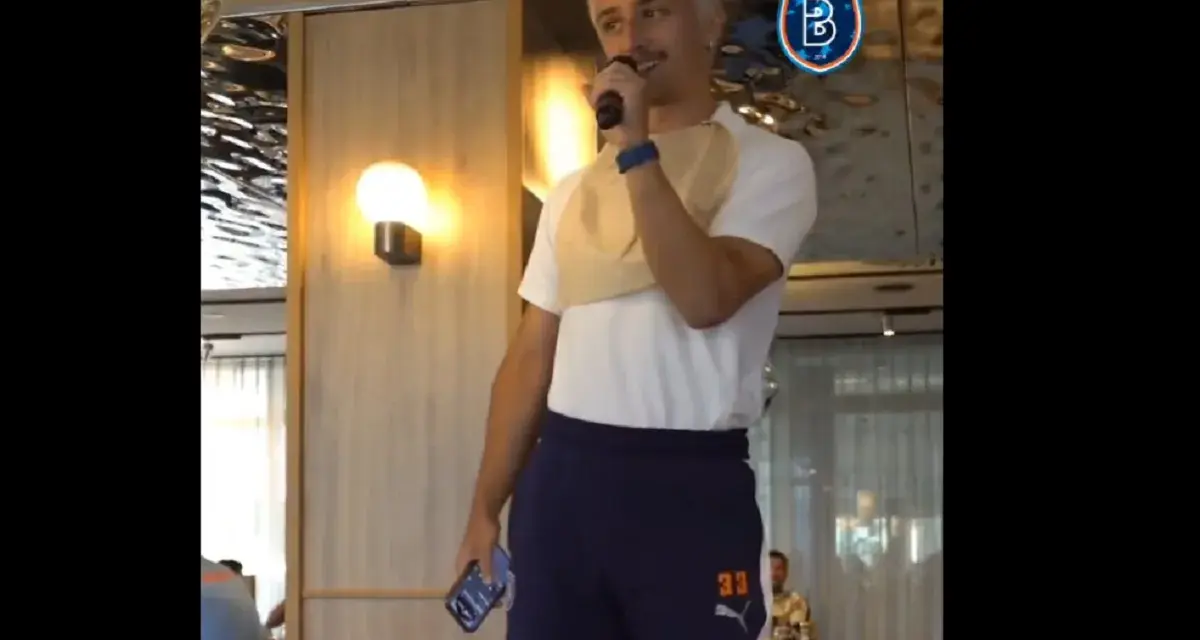 Młody mężczyzna z blond włosami, w koszulce i granatowych spodenkach z numerem 33, trzyma mikrofon. W tle logo İstanbul Başakşehir.