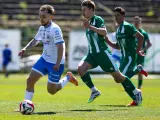 Piłkarz w białej koszulce z niebieskimi spodenkami prowadzi piłkę na boisku. W tle zawodnicy w zielonych strojach. Mecz forbet 4 liga warmińsko-mazurska.