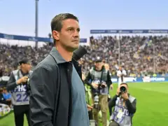 Trener Interu wpatrzony w murawę, otoczony przez fotoreporterów na tle trybun pełnych kibiców.