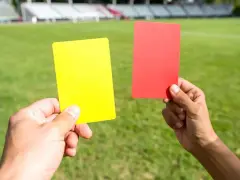 Dwie ręce trzymają żółtą i czerwoną kartkę na tle boiska piłkarskiego. Sędzia pokazuje czerwoną kartkę w piłce nożnej.