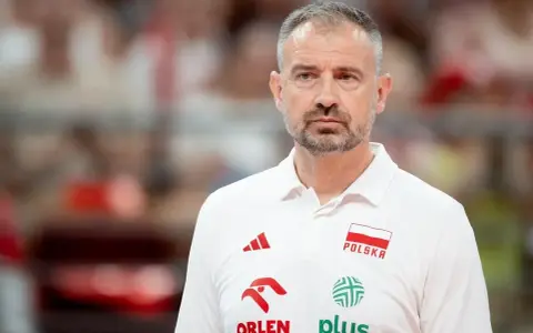 Skupiony trener siatkarzy reprezentacji Polski w białej koszulce z godłem narodowym i logotypami sponsorów.