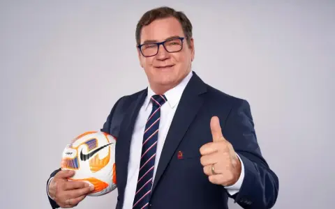 Uśmiechnięty trener reprezentacji Polski w garniturze trzyma piłkę i pokazuje kciuk w górę.