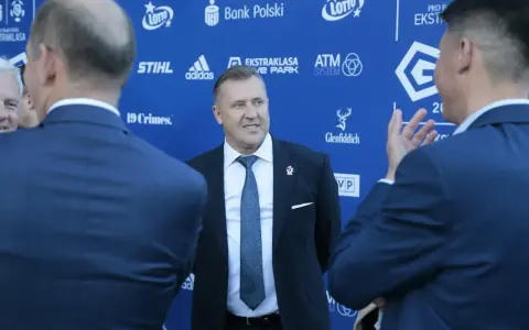 Mężczyzna w garniturze uśmiecha się, stojąc na tle niebieskiej ściany z logotypami sponsorów, być może rozmawiając o tym, ile dostają kluby ekstraklasy.