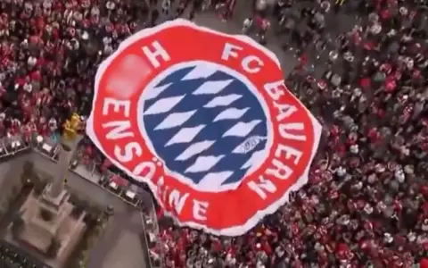 Ogromna flaga z herbem FC Bayern Monachium niesiona przez tłum kibiców.