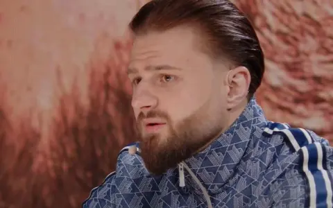 Amadeusz Ferrari w niebieskiej bluzie z białymi paskami, z charakterystyczną fryzurą i brodą, siedzi przed ekranem.