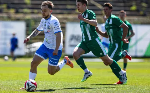 Piłkarz w białej koszulce z niebieskimi spodenkami prowadzi piłkę na boisku. W tle zawodnicy w zielonych strojach. Mecz forbet 4 liga warmińsko-mazurska.