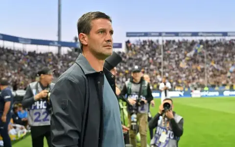 Trener Interu wpatrzony w murawę, otoczony przez fotoreporterów na tle trybun pełnych kibiców.