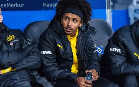 Piłkarz Adeyemi w czarnej kurtce z żółtym logo BVB i napisem REWE, siedzi na ławce rezerwowych, trzymając butelkę z napojem.
