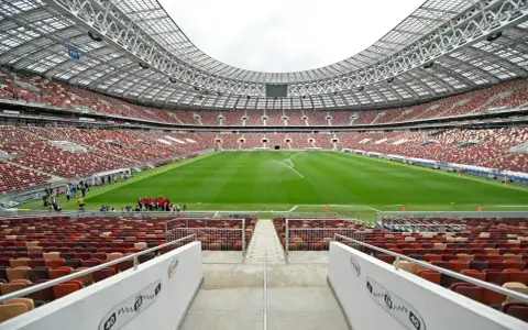 Pusta trybuna moskiewskiego stadionu z rzędem czerwonych i brązowych siedzeń. Na boisku pracują zraszacze.