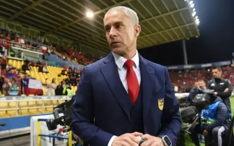 Sylvinho, trener w granatowym garniturze i czerwonym krawacie, stoi na tle trybun stadionu z flagą Chile.