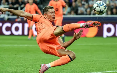Piłkarz w pomarańczowym stroju wykonuje akrobatyczne kopnięcie do piłki. To Robert Lewandowski, który gra w Lidze Mistrzów.