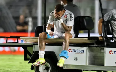 Neymar siedzi na wózku golfowym, zakrywając twarz dłońmi. Jego kontuzja jest widoczna, a zawodnik wygląda na zrozpaczonego.