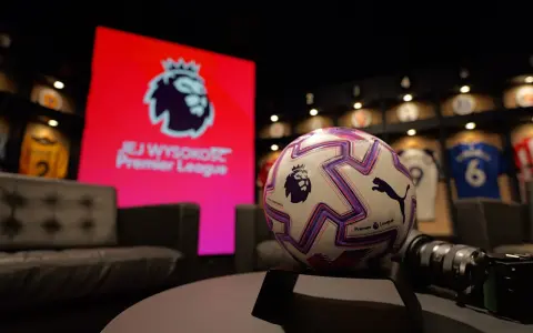Piłka do gry w Premier League na tle ekranu z logo ligi. W tle widoczne koszulki piłkarskie.
