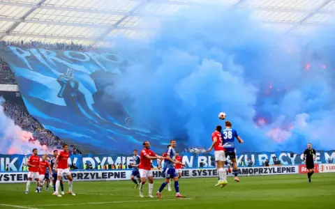 Mecz Ruch Chorzów – Wisła Kraków. Niebieskie dymy i ogromna oprawa kibiców na trybunach. Piłkarze walczą o piłkę na murawie.
