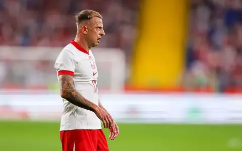 Kamil Grosicki w białej koszulce z orzełkiem i czerwonych spodenkach, gotowy do gry. Jego obecne drużyny to Polska i Pogoń Szczecin.