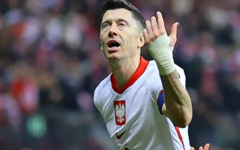 Robert Lewandowski w koszulce reprezentacji Polski, z bandażem na ręce, wznosi gest ku górze. Fani zastanawiają się, o której mecz piłki nożnej.