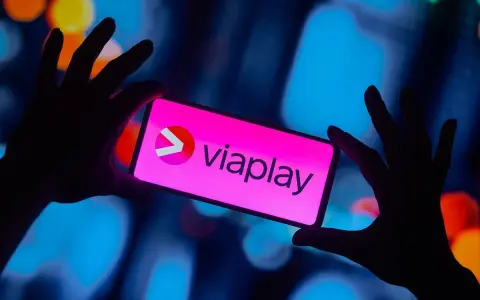 Dłonie trzymają smartfon z logo Viaplay. W tle rozmyte, kolorowe światła.