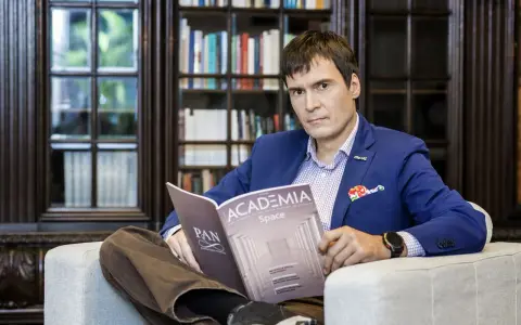 Mężczyzna w garniturze czyta magazyn "Academia Space" w bibliotece. W tle regały z książkami.